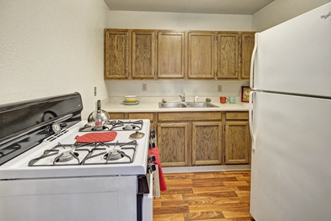 Cozy kitchen here at the Village at Calais Apartments with wooden cabinets, a gas stove, and white fridge. Counter features a double sink and colorful mugs, conveying a warm, homely feel.