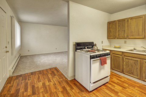 Small kitchen the Village at Calais Apartments with wooden cabinets, white stove, and wooden flooring. Adjacent is a carpeted living area; both areas are empty, creating a neutral, simple tone.