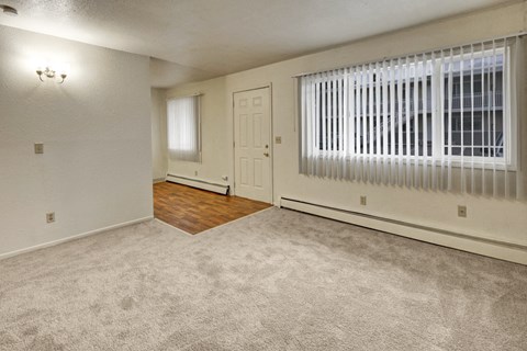 A vacant living room here at Village at Calais Apartments with light beige walls and carpet, featuring a large window with vertical blinds. The entry area has hardwood flooring.