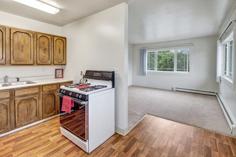Compact kitchen here at the Village at Calais Apartments with wood cabinets, white stove, and a red towel, opens to a carpeted living room with large window showing lush greenery. Bright and cozy.