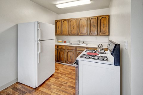 Small kitchen here at the Village at Calais Apartments with wood cabinets, white fridge, and gas stove. Wooden floor, overhead light. Cozy and simple, with a pot on the stove.
