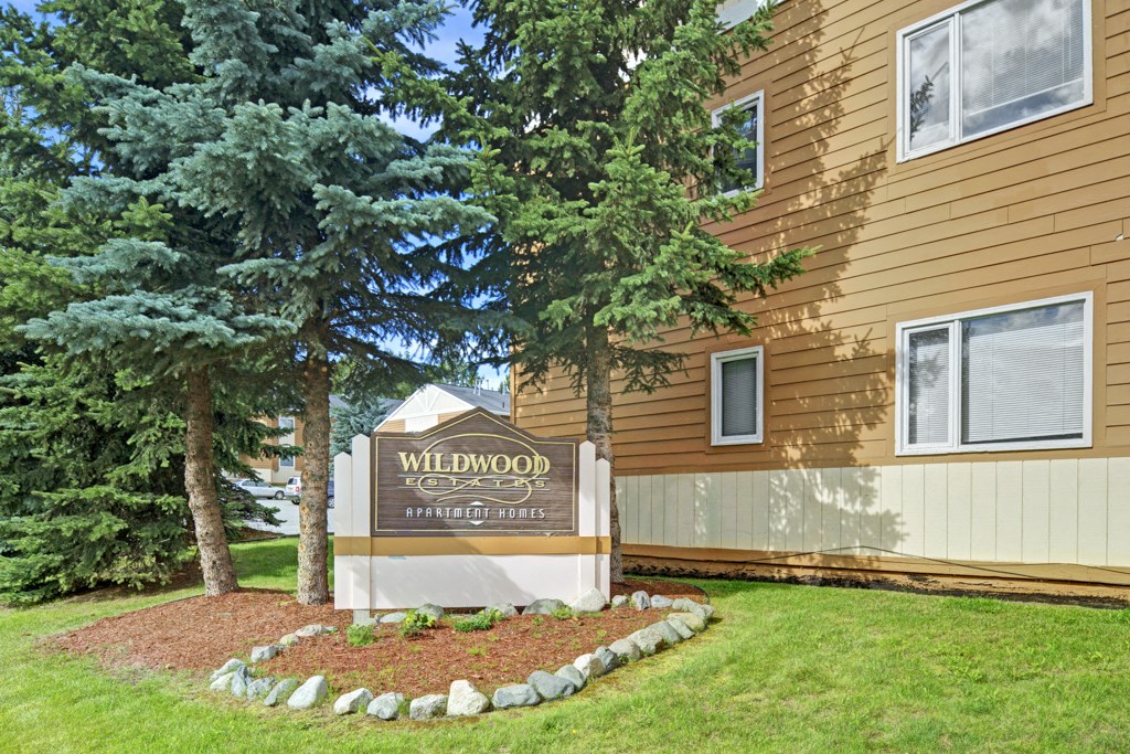 Sign for Wildwood Estates Apartment Homes, surrounded by lush green grass and tall pine trees, next to a tan building with multiple windows.