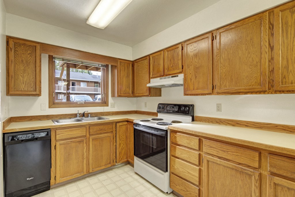 A kitchen here at Wildwood Estates Apartments with wooden cabinets, a stove, and dishwasher. A window above the sink overlooks a parking area. Bright and clean ambiance.