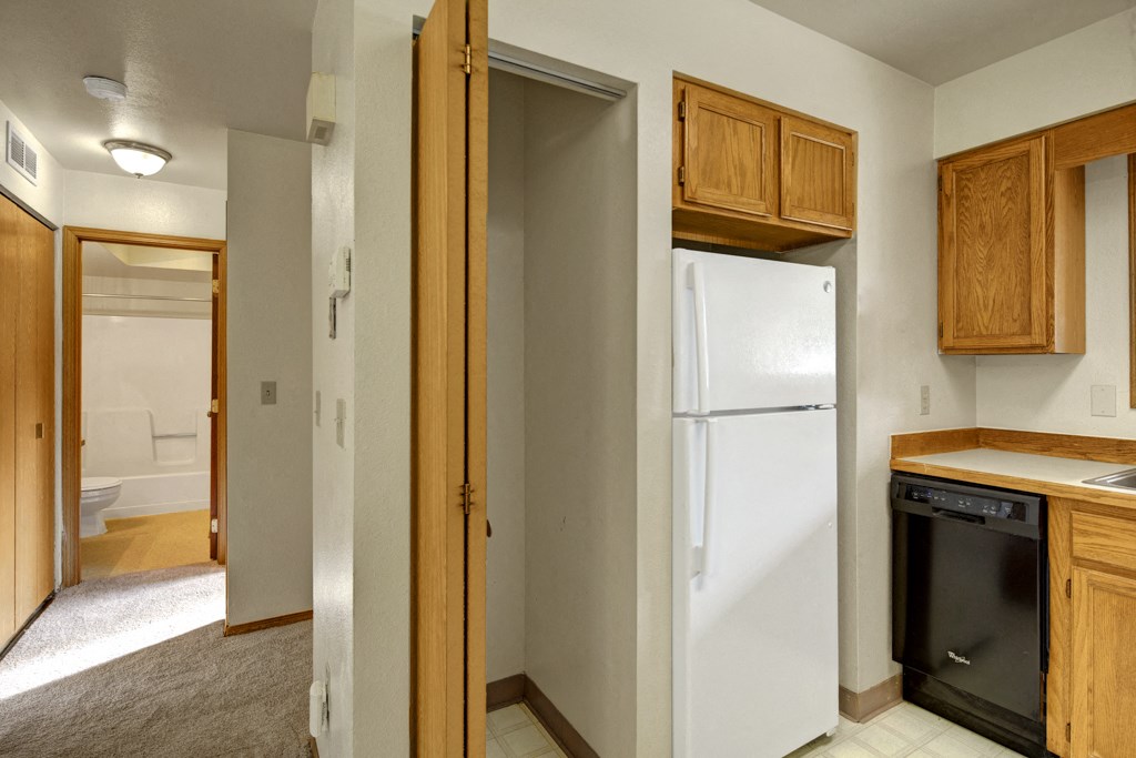 A kitchen here at Wildwood Estates Apartments with wooden cabinets, a white refrigerator, and a black dishwasher. Adjacent hallway leading to a bathroom, with a warm and clean ambiance.