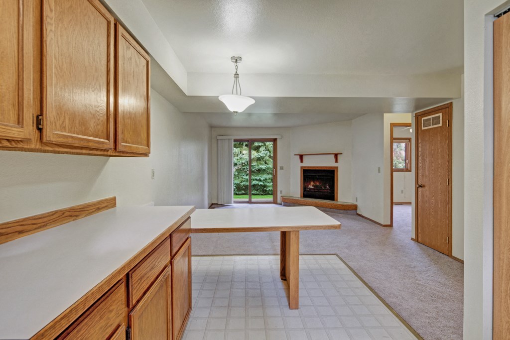 Spacious, bright interior here at Wildwood Estates Apartments with light wood cabinets, tiled floors transitioning to carpet. Features fireplace, sliding door to greenery, modern yet cozy feel.