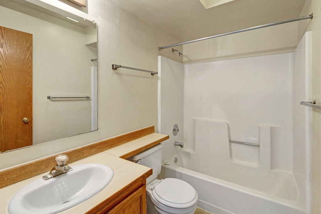 Small, tidy bathroom here at Wildwood Estates Apartments with white fixtures. A sink with wooden cabinet, toilet, and bathtub with shower inside are visible. Neutral color tones.