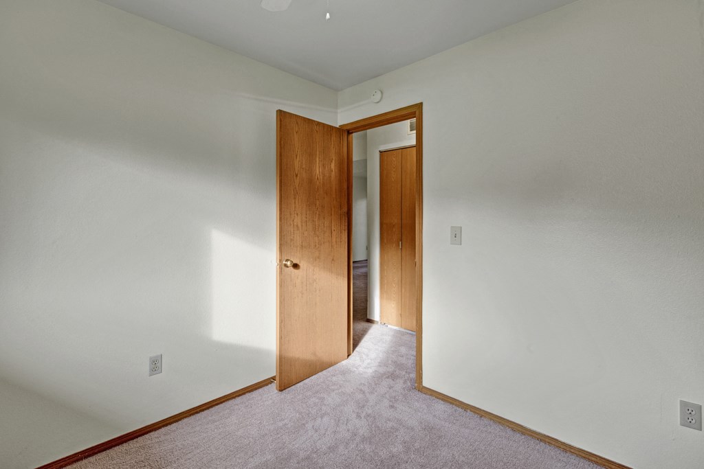 Empty bedroom here at Wildwood Estates Apartments with off-white walls, beige carpet, and an open wooden door. Sunlight casts soft shadows, creating a calm and inviting atmosphere.