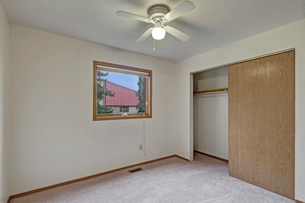 Plain bedroom here at Wildwood Estates Apartments with beige carpeting and white walls, featuring a ceiling fan. A window shows a view of a tree and red-roofed building. Open closet with wooden sliding door.