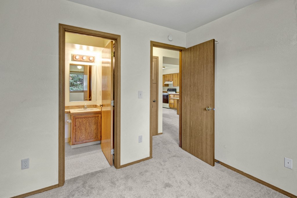 A beige carpeted room here at Wildwood Estates Apartments with two doors; one leads to a lit bathroom featuring a wooden vanity, and the other to a kitchen with wooden cabinets.