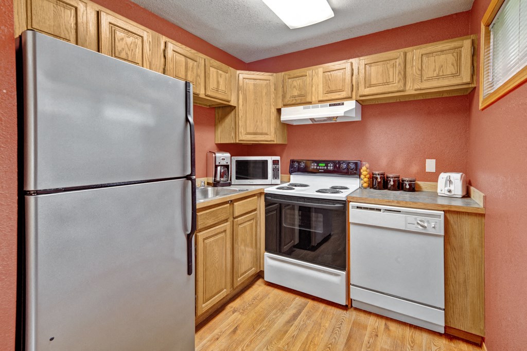 Compact kitchen here at Wildwood Estates Apartments with wooden cabinets and red walls. Features a stainless steel fridge, stove, microwave, dishwasher, toaster, jars, and wooden floor. Cozy atmosphere.