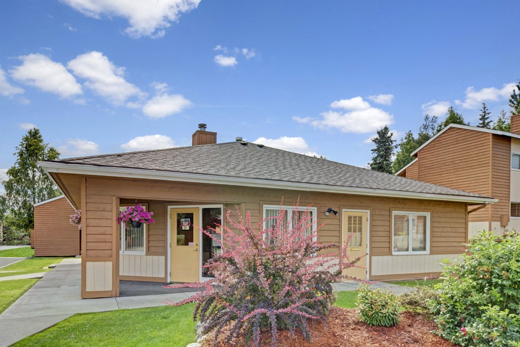 A single-story, brown wooden house here at Wildwood Estates Apartments with a grey roof sits under a bright blue sky. Pink flowers and green shrubs adorn the front, adding a welcoming touch.