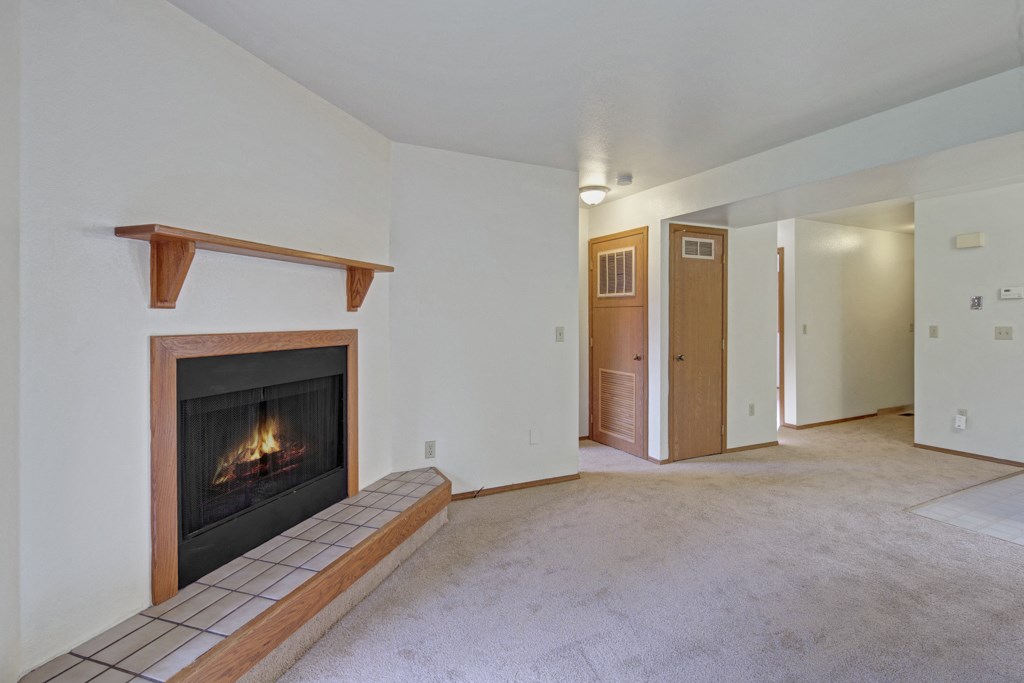 Cozy living room here at Wildwood Estates Apartments with a lit fireplace framed by wood, beige carpet, and white walls. Soft lighting from a wall lamp adds warmth to the space.