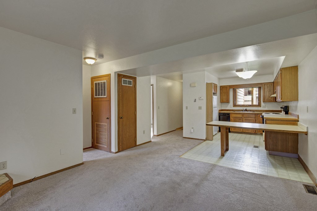 Spacious, empty living area here at Wildwood Estates Apartments with beige carpet, wood panel doors, and an adjoining kitchen. The kitchen features wooden cabinets and vinyl flooring, creating a simple, neutral tone.