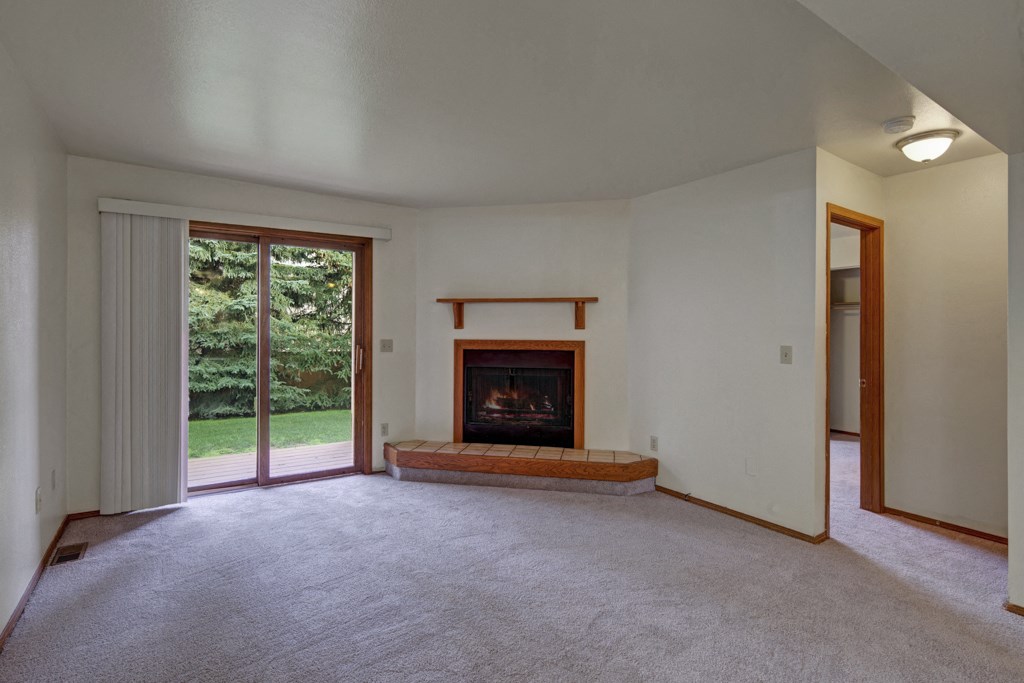 Living room here at Wildwood Estates Apartments with a cozy fireplace, white walls, grey carpet, and a sliding glass door opening to a lush green garden, creating a serene atmosphere.