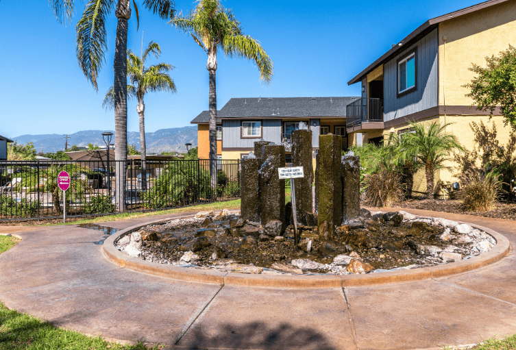 A small fountain in the middle of a circular walkway with a sign in front of a building at Raintree Apartments, CA 92346