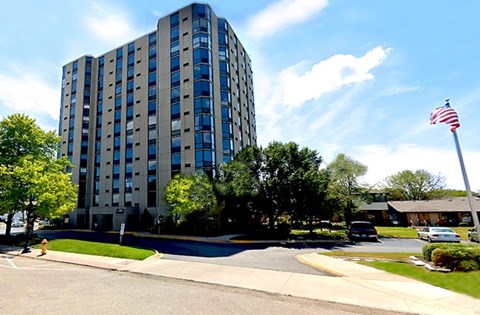 A tall building with a flag on top is surrounded by trees and a street in front.