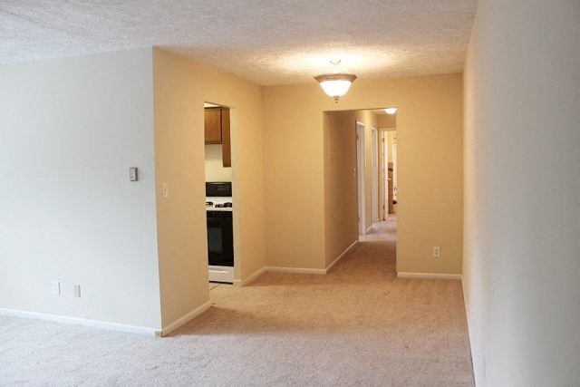 an empty living room with a kitchen and an open hallway