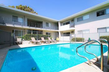 A swimming pool in front of a white and pink building with lounge chairs.