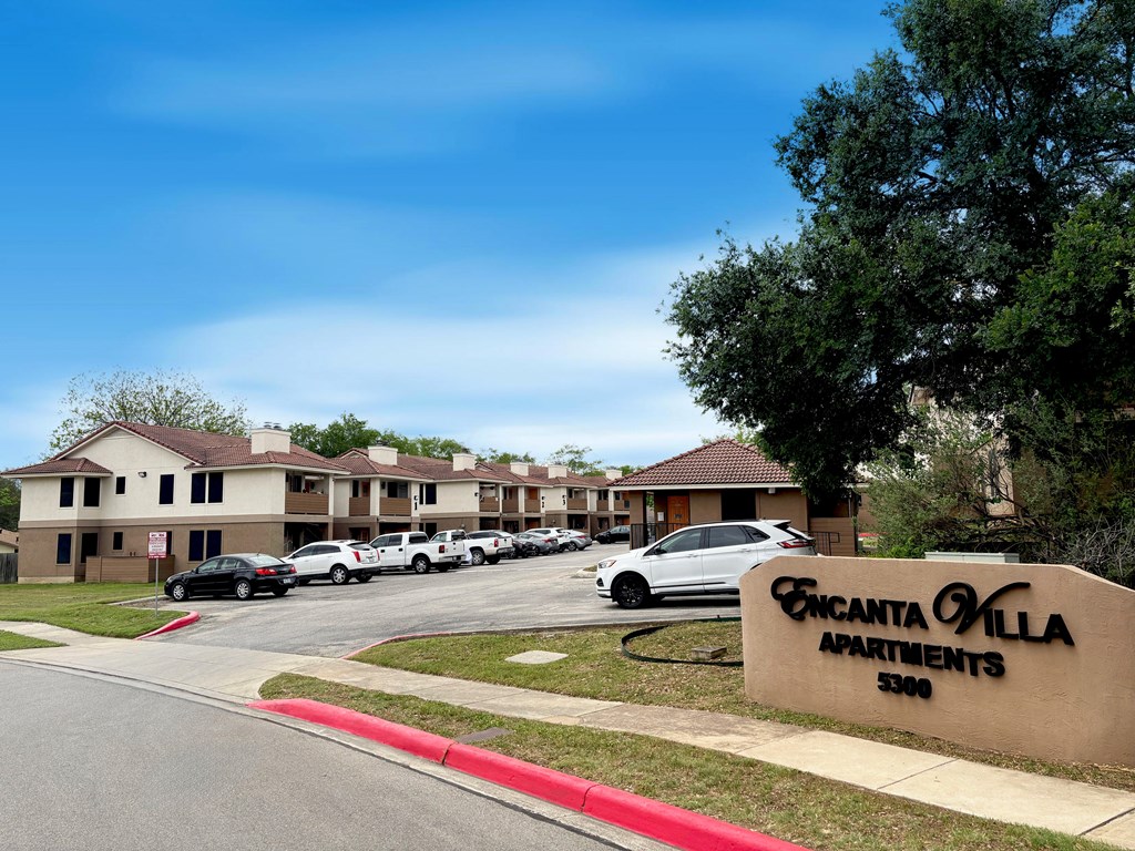 A sign for Encanta Villa Apartments is in front of a row of parked cars.