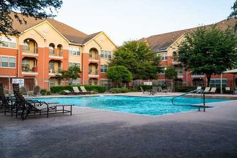 A large swimming pool is surrounded by lounge chairs and trees in front of apartment buildings.