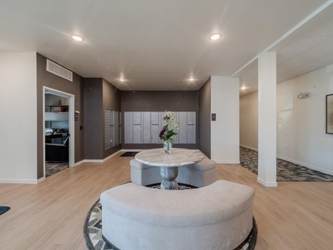 A round grey ottoman sits in the middle of a spacious room with a white table in front of it.