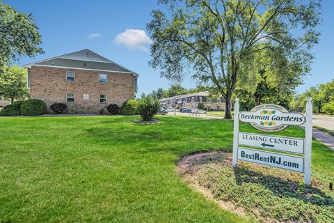 Exterior photo of Beekman Gardens in Hillsborough, NJ showing the entry sign, and a road leading to several apartment buildings. Lush green landscaping and trees can be seen around the property.