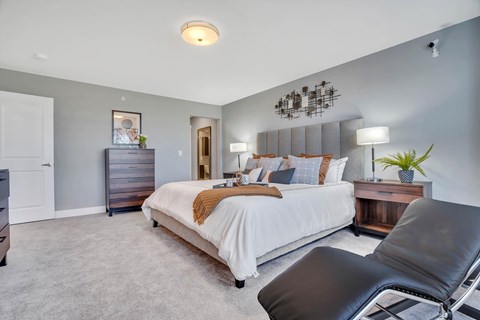 Interior photo of the Primary Bedroom suite in a typical apartment at Ewing Town Center showing a bed, several dressers, end table with lamp, a lounge chair. There is a doorway showing access to the primary bath