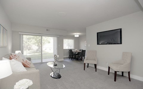 A living room with a grey couch, a coffee table, a dining table with chairs, and a flat screen TV mounted on the wall.