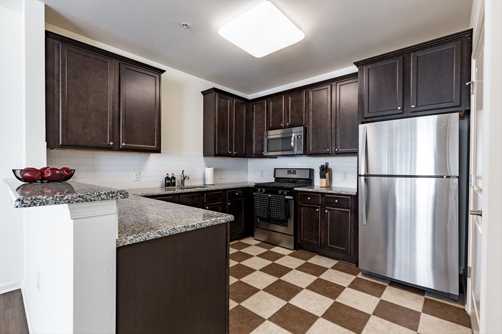 Modern kitchen at Queens Gate Apartments featuring stainless steel appliances, dark wood cabinetry, granite countertops, and a checkered tile floor.