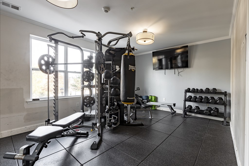 Fully equipped fitness center at Queens Gate Apartments featuring weight training equipment, a punching bag, dumbbells, and a flat-screen TV for an enhanced workout experience.
