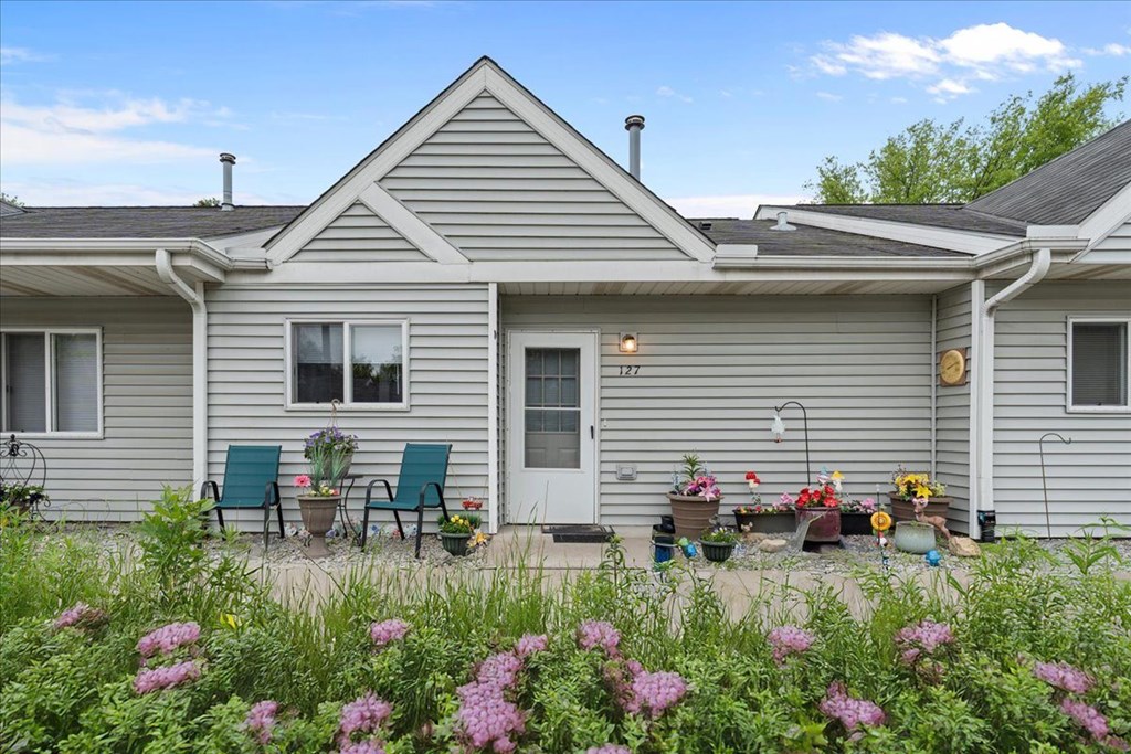 A house with a grey exterior and a white door is surrounded by a garden with pink flowers.