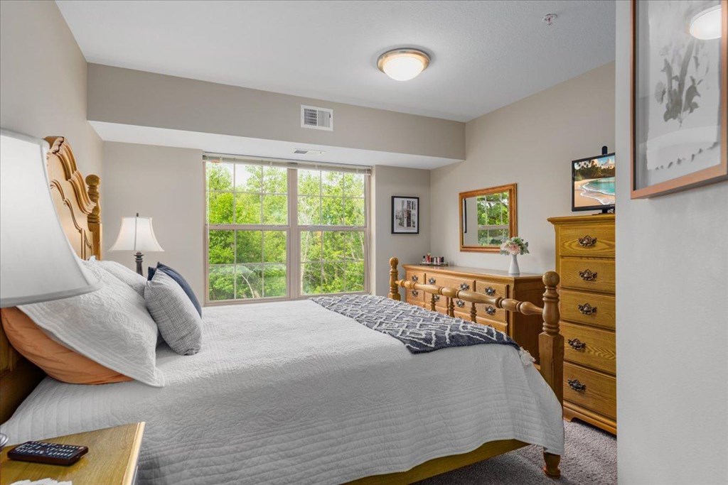 A bedroom with a large bed, a dresser, and a window overlooking a green area.