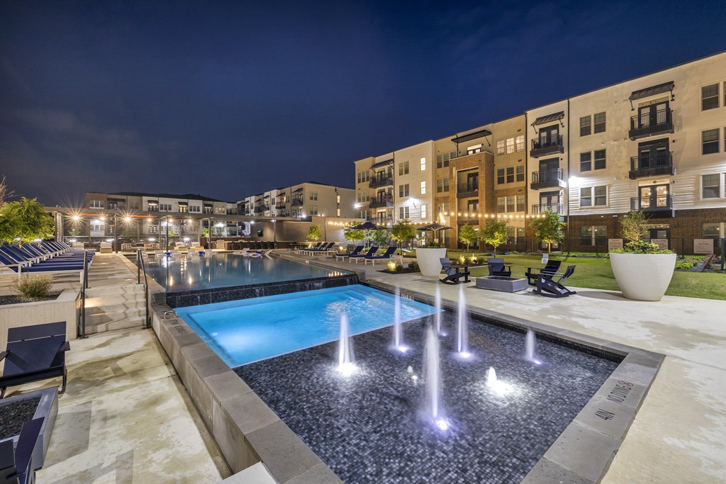 a pool with water fountains in front of an apartment building at night