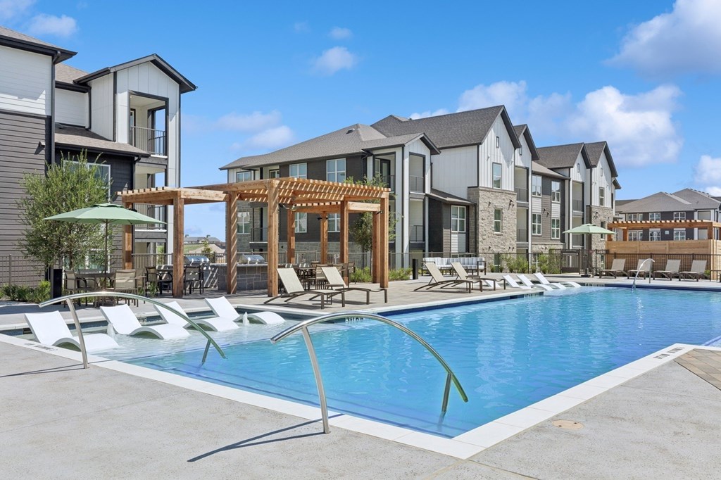 A swimming pool surrounded by lounge chairs and umbrellas in front of apartment buildings.