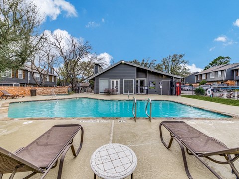 A pool with lounge chairs and a table in the foreground with a building in the background.