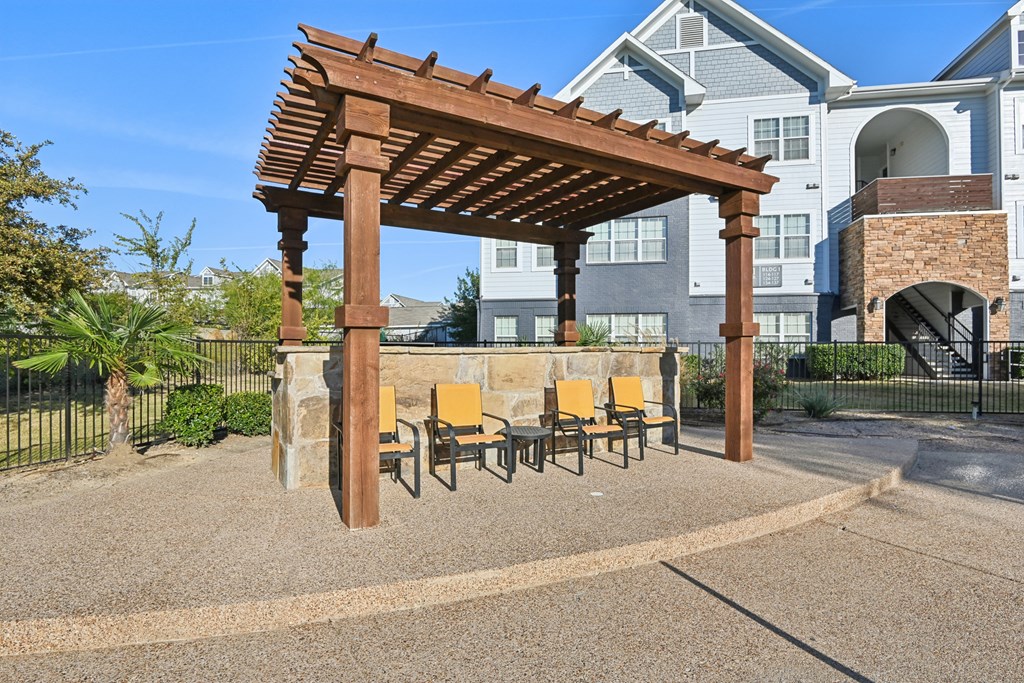 A wooden pergola with yellow chairs is in the foreground of a residential area.