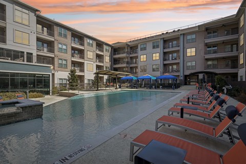 A swimming pool surrounded by sun loungers and umbrellas in front of apartment buildings.