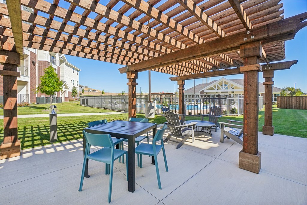 A wooden pergola with a table and chairs is in the foreground of a patio.