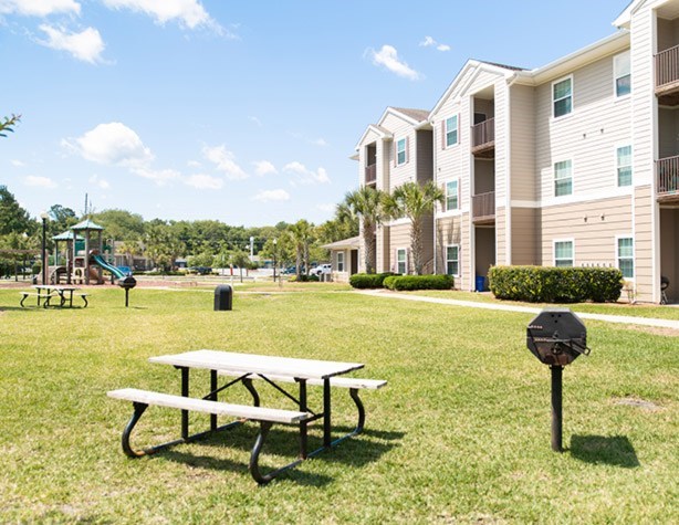 A picnic table is in the foreground of a grassy area with apartment buildings in the background.