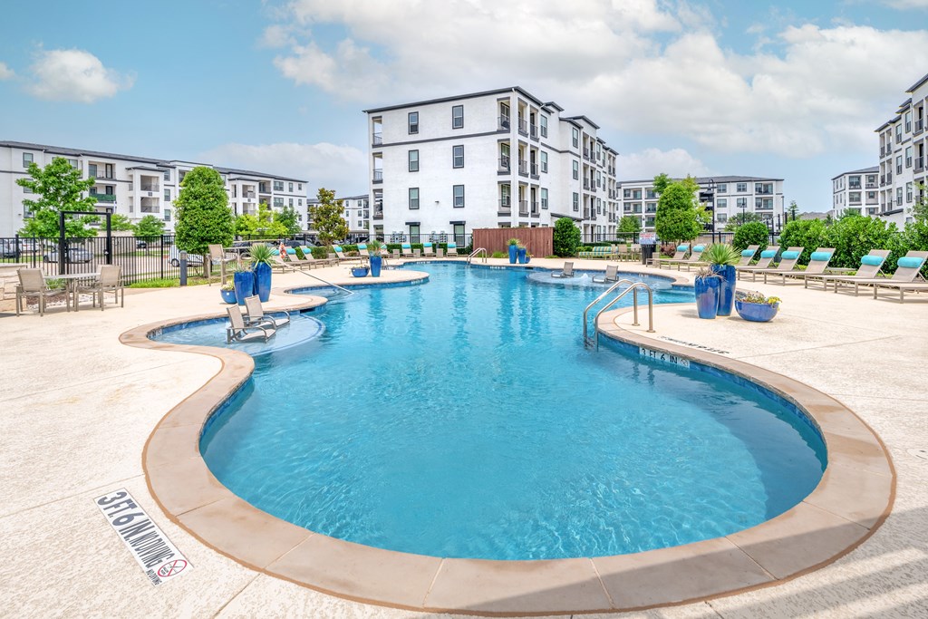 A large outdoor swimming pool surrounded by lounge chairs and buildings in the background.