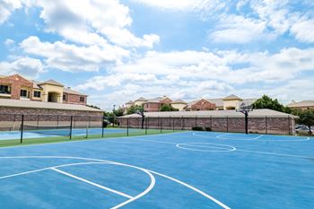 A basketball court with a blue surface and white lines is surrounded by a brick wall and houses.