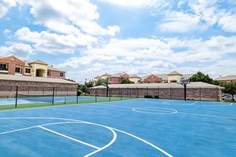 A basketball court with a blue surface and white lines is surrounded by a brick wall and houses.