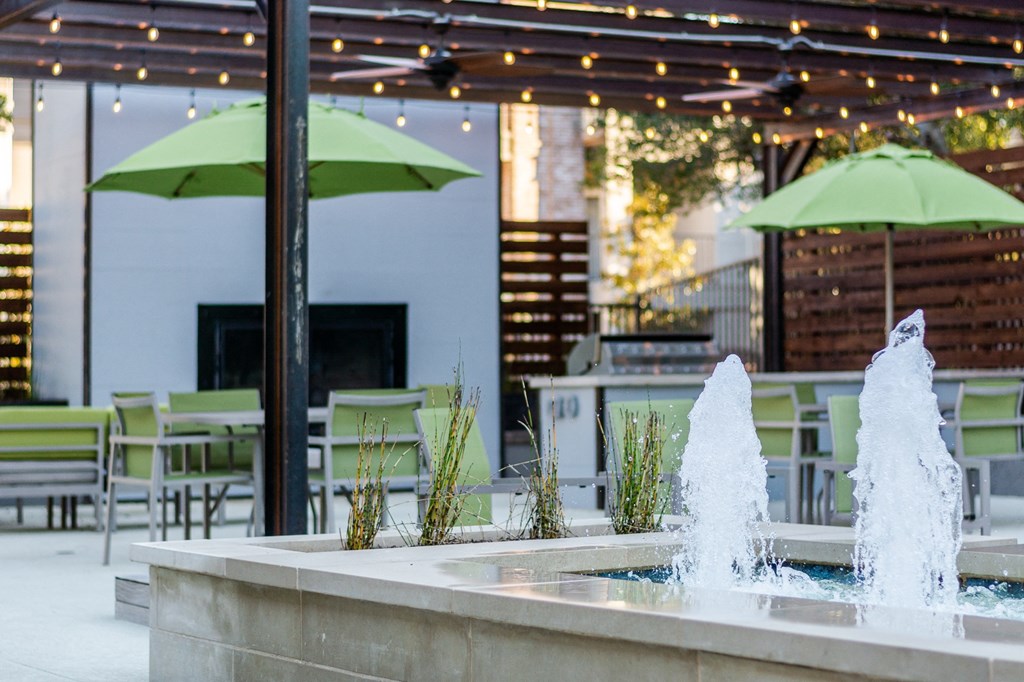a fountain in the middle of a patio with green chairs and umbrellas in the background