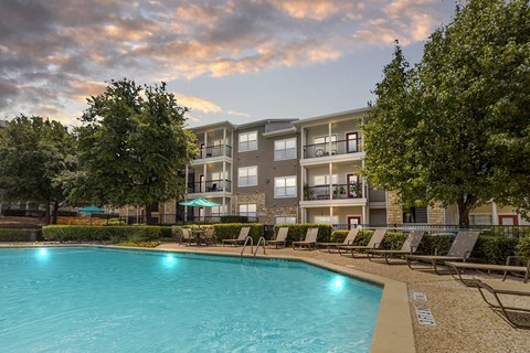 A large swimming pool is surrounded by lounge chairs and trees in front of apartment buildings.