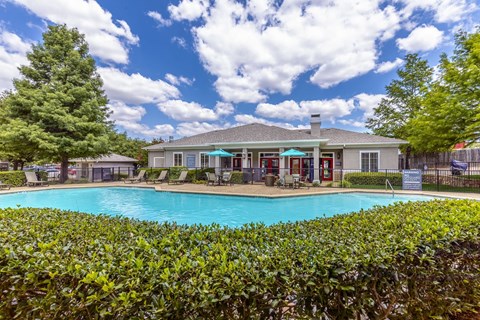 A pool surrounded by green bushes with a house in the background.
