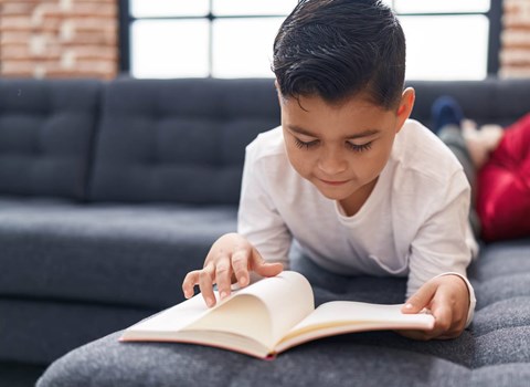 A young boy is reading a book on a grey couch.