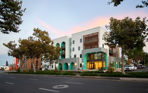 A modern building with a white facade and green accents is surrounded by trees and parked cars.