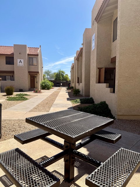A black table with four benches is in the foreground of a sunny courtyard.