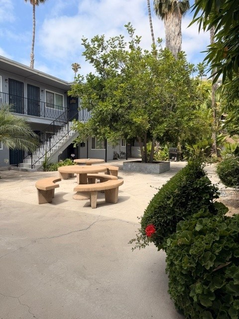 A concrete patio with a table and chairs surrounded by greenery and a building in the background.