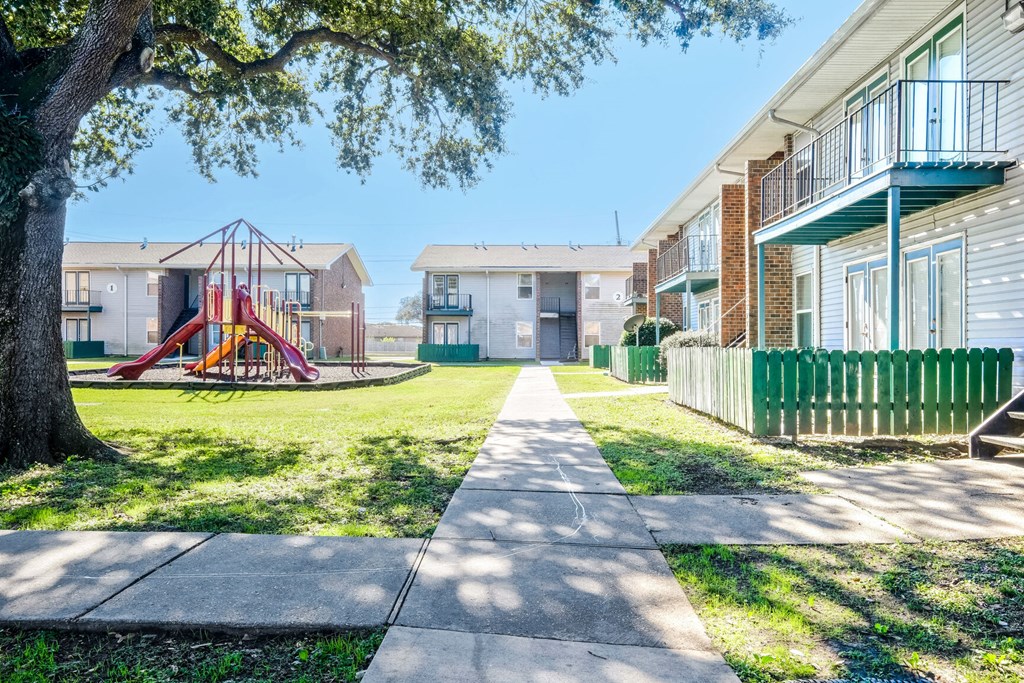 A playground with a slide is in the middle of a grassy area between two apartment buildings.